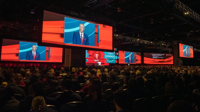 Crowded auditorium with multiple screens featuring speaker