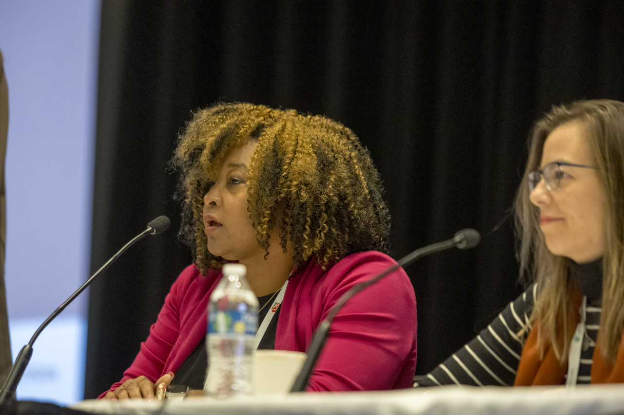 Two women speaking at a conference panel with microphones and water bottles