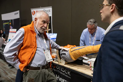 Man in orange vest demonstrates product at HomeShock trade show booth while visitor examines yellow gloved hand model