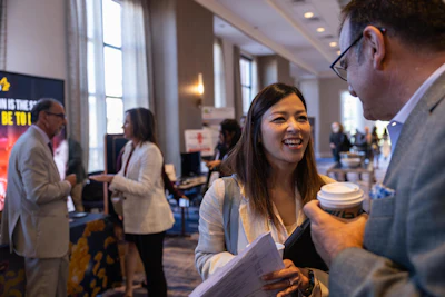 Professional woman smiling while holding coffee and documents during a corporate networking event with colleagues in background