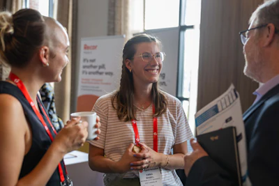 Three professionals wearing conference badges engage in conversation at a business event near a window.