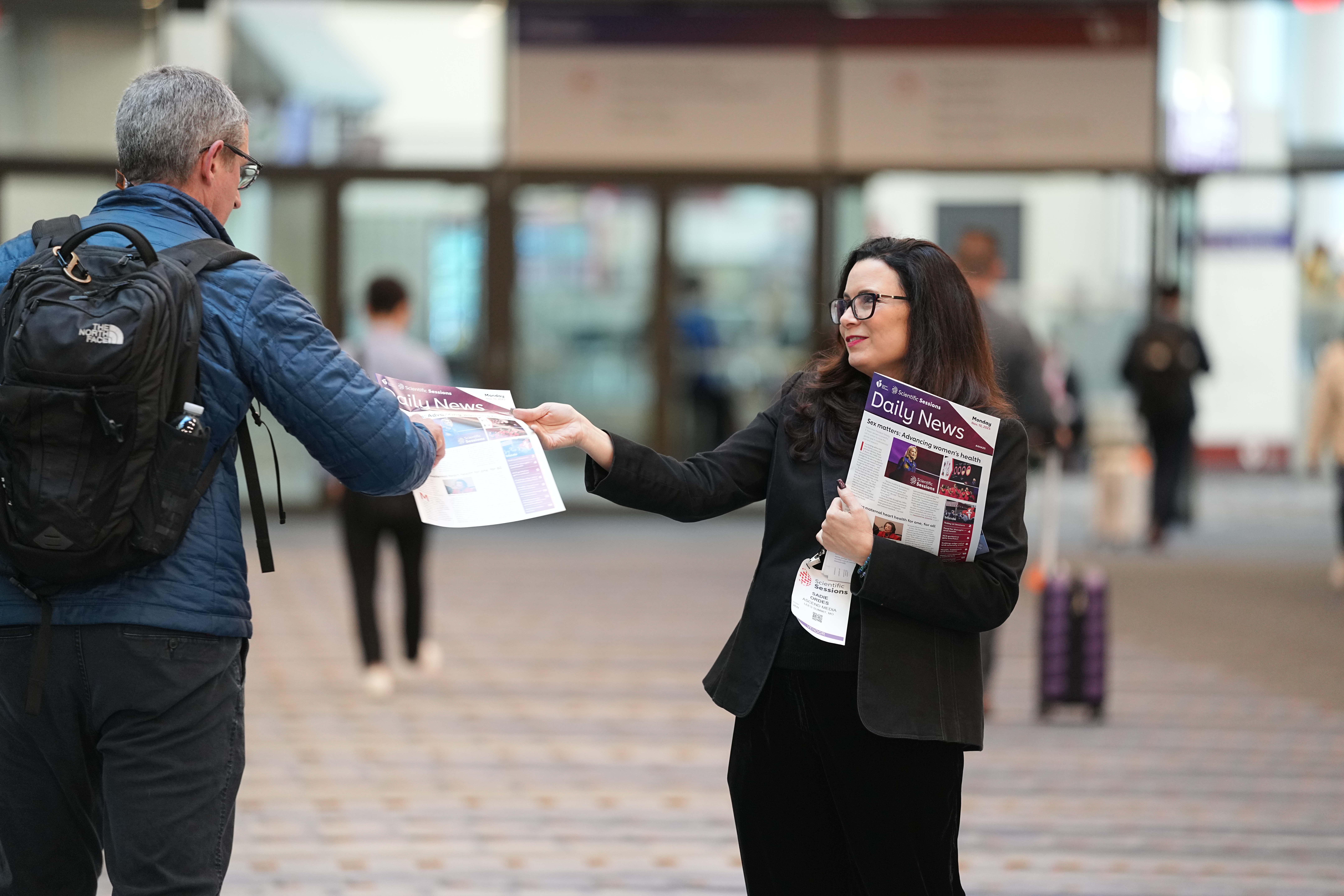 Woman in black blazer distributing newspaper to man wearing blue jacket and North Face backpack in urban transit station