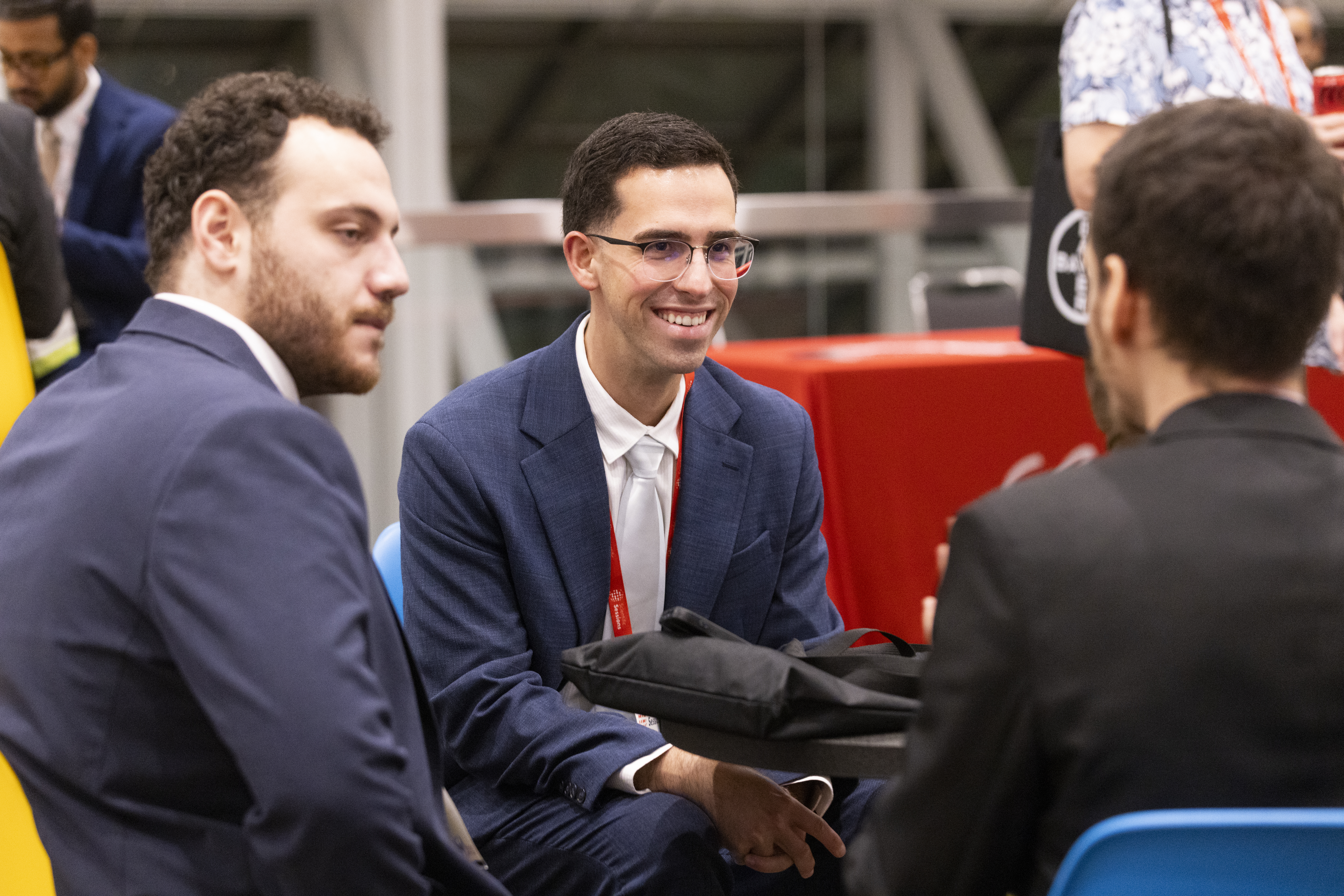 Three men in business suits conversing at an indoor event, with one wearing glasses and smiling