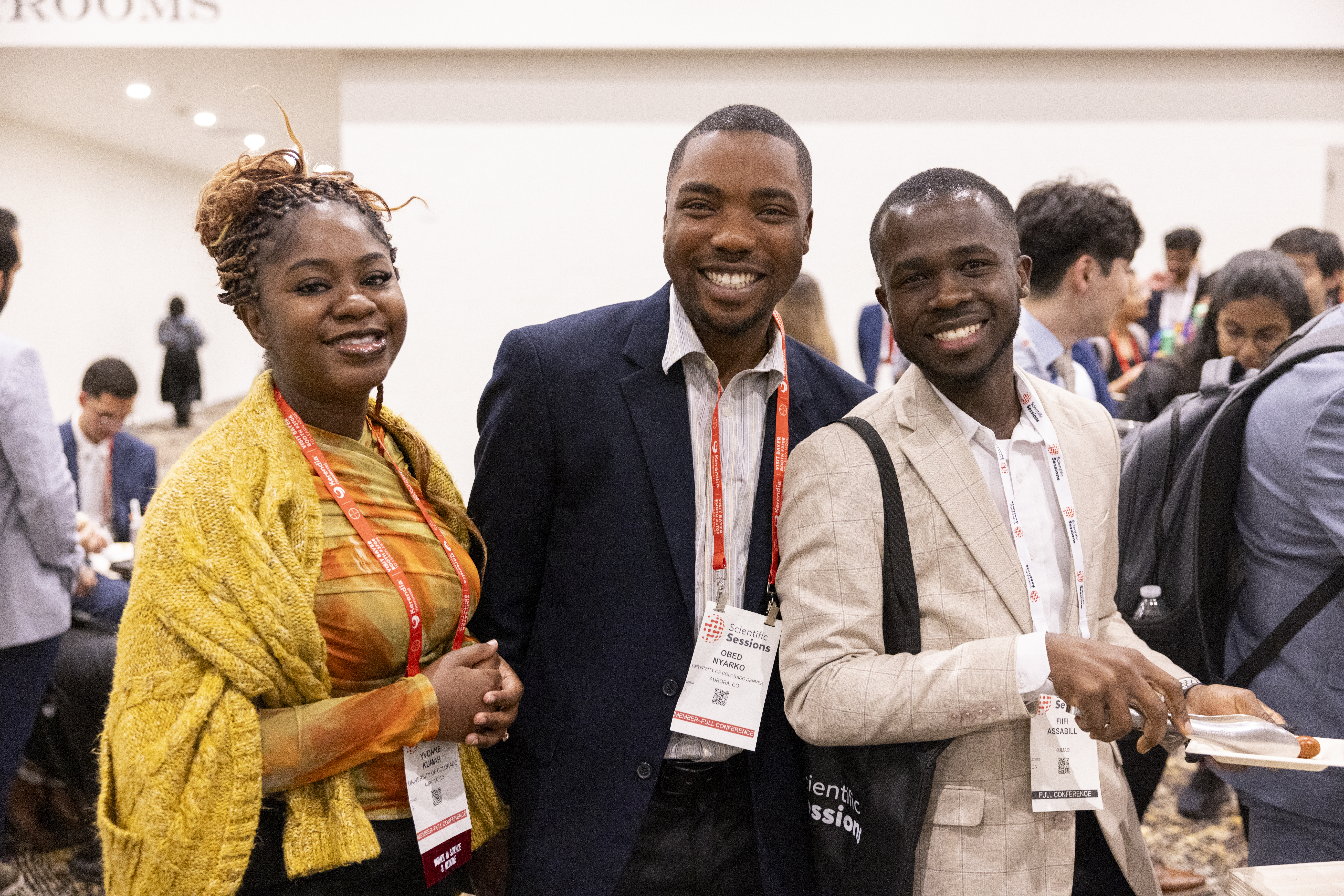 Three professionals smiling at a conference event, wearing credential badges in an indoor venue with other attendees visible in background.