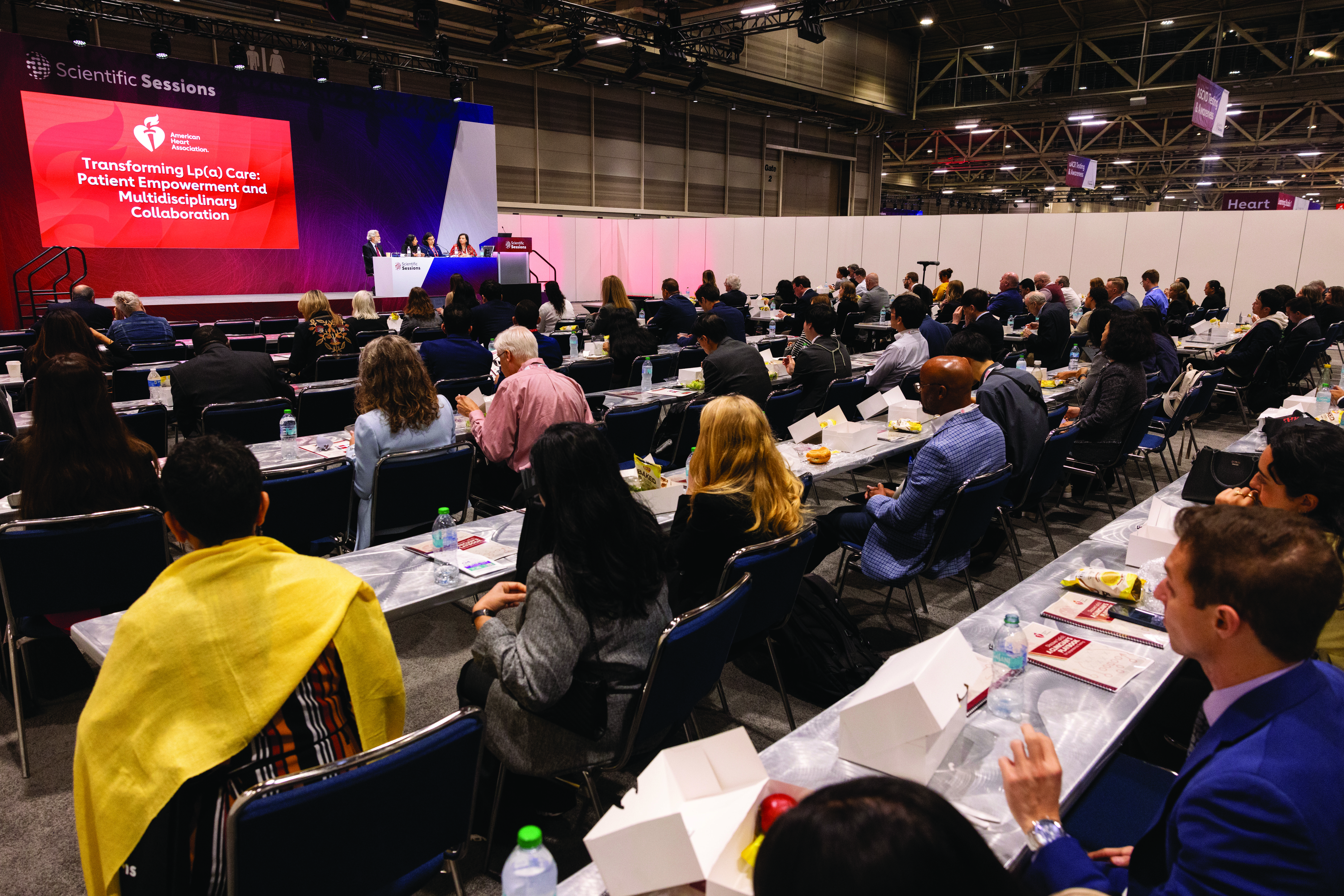 Large conference hall with attendees seated at tables facing a stage with projection screen displaying scientific session content.