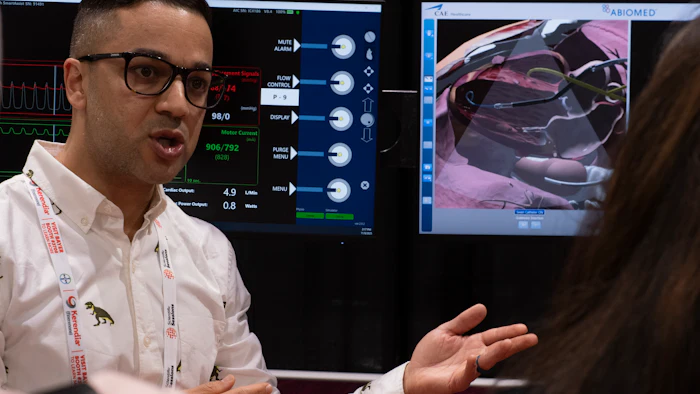 Professional presenter wearing glasses and white shirt gestures while speaking at tech event with monitoring displays in background