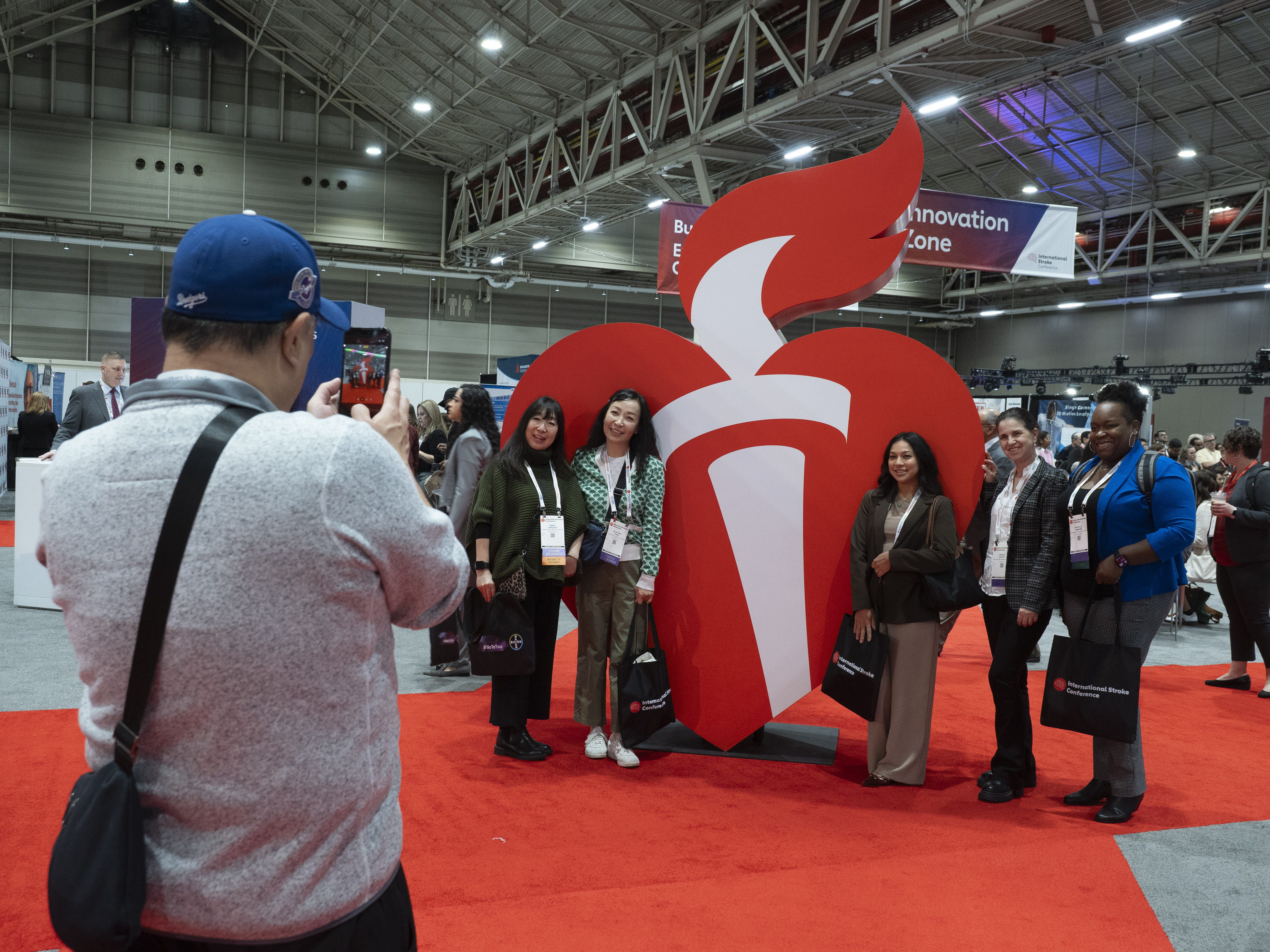 Group of attendees posing for photos at a trade show booth with a large red and white 3D logo sculpture on an orange floor.