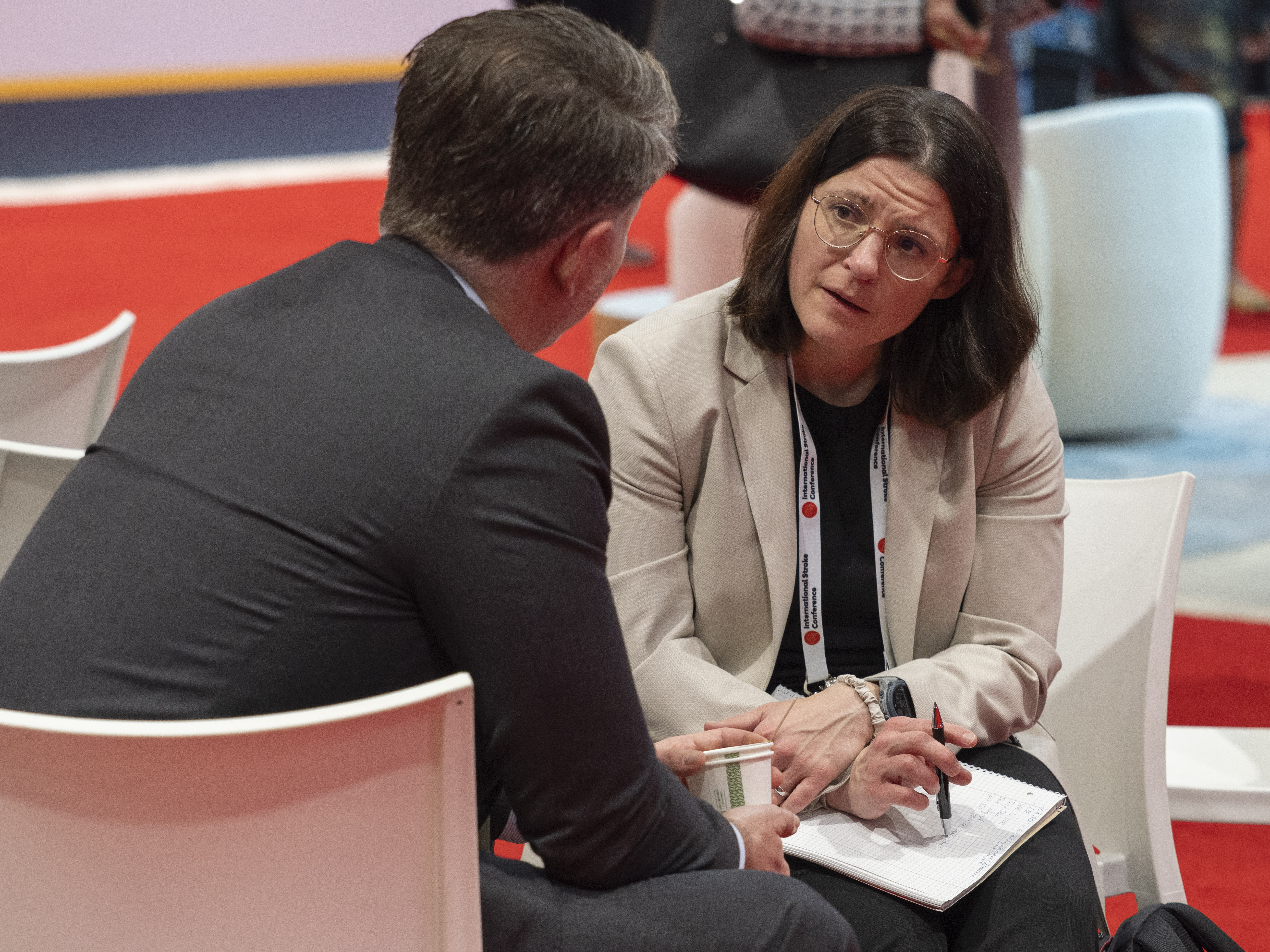 Two business professionals in conversation at a corporate conference, with woman in beige blazer taking notes.