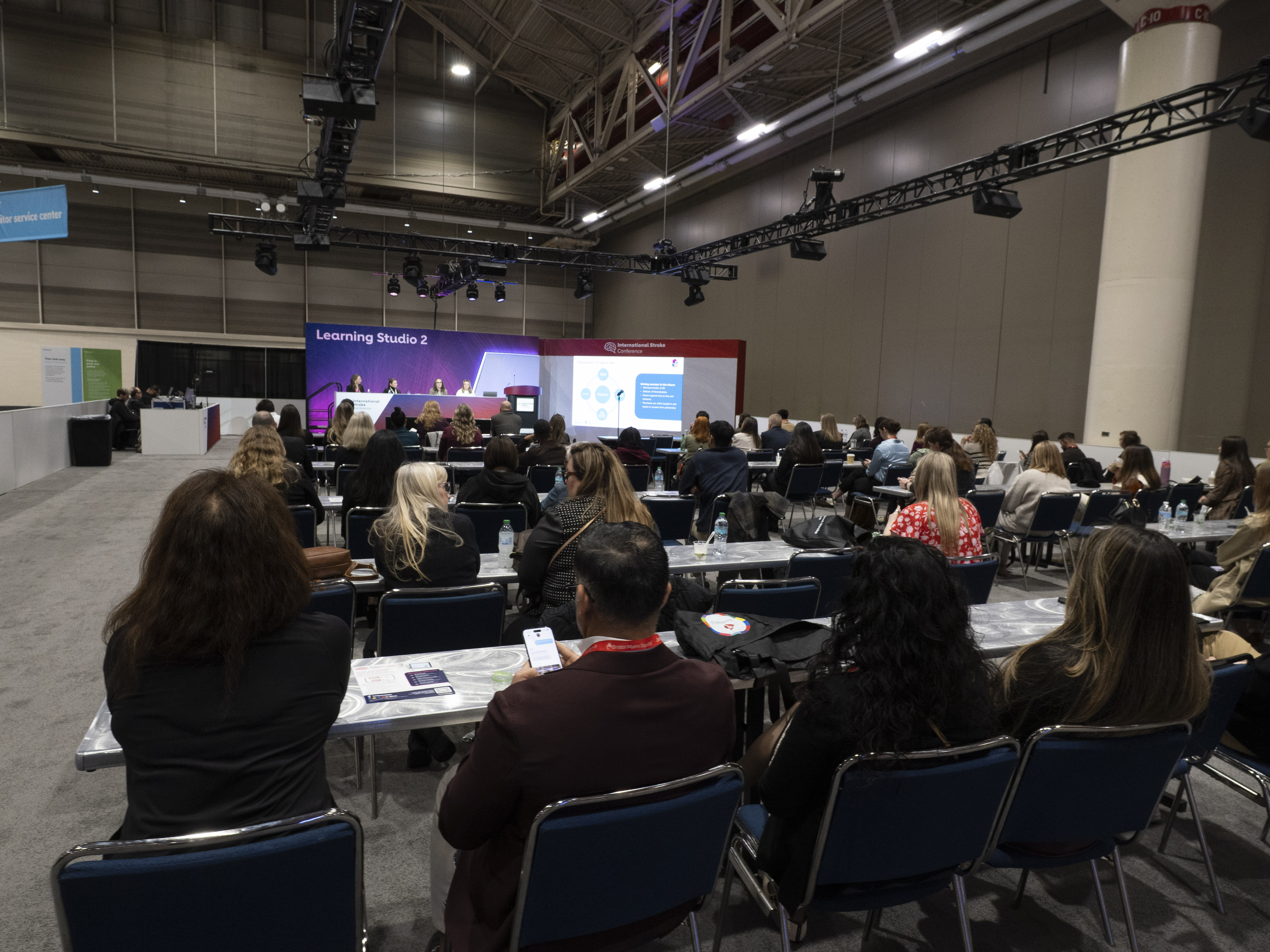 Large conference room with attendees seated at tables facing presentation screens in Learning Studio 2