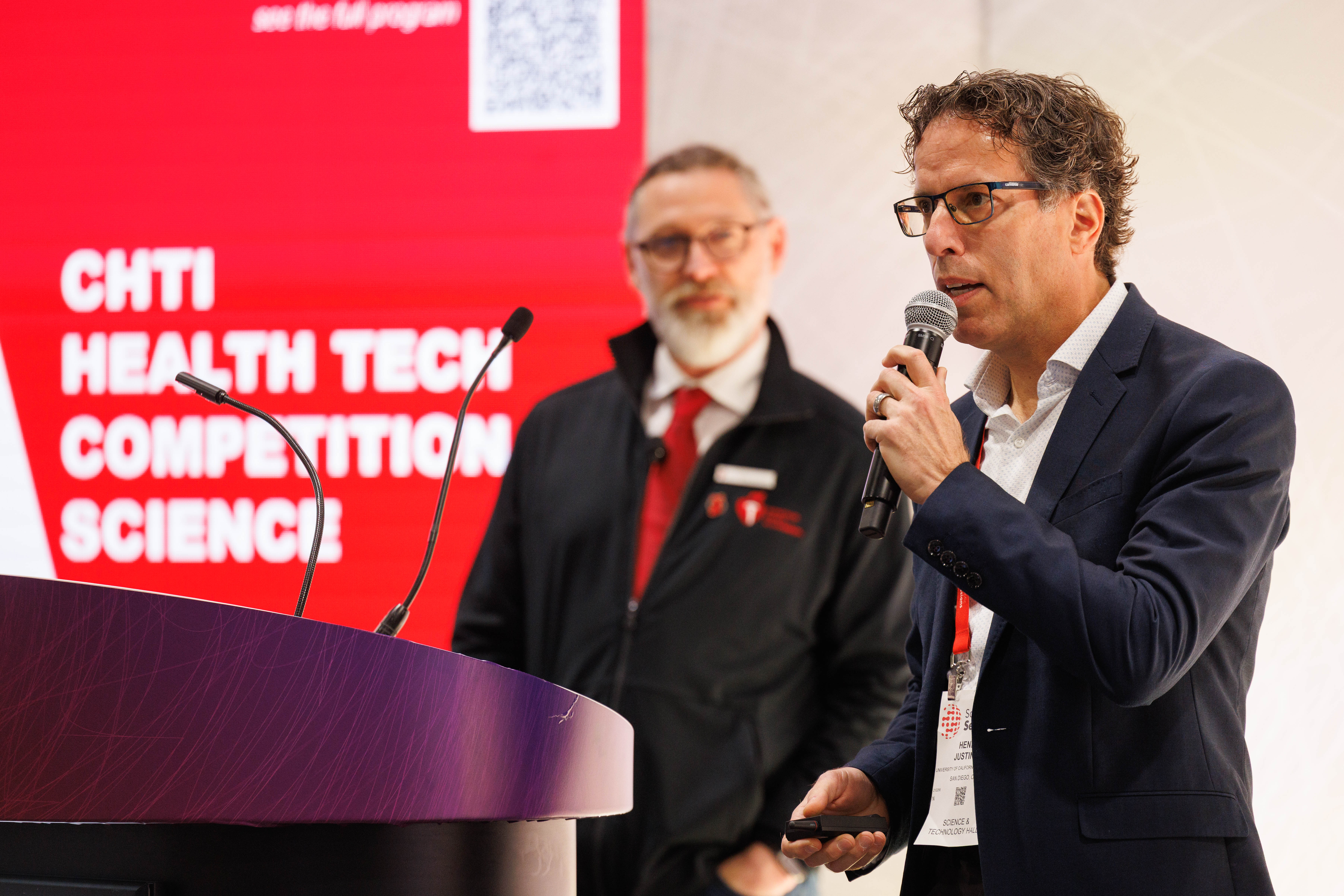 Man speaking at podium during CHTI Health Tech Competition Science event with colleague in background
