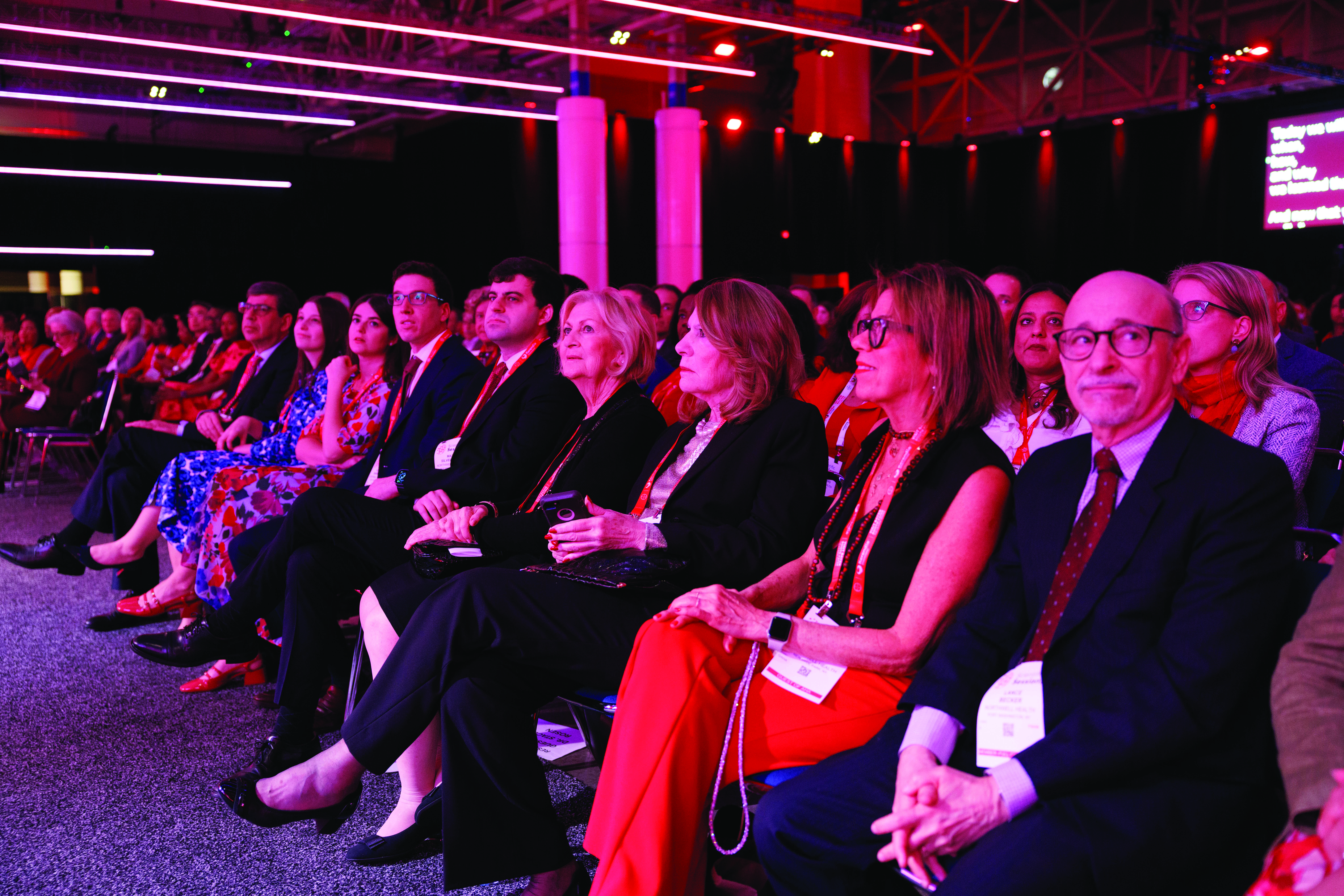 Audience members in formal attire seated in an auditorium with pink accent lighting during a corporate conference or formal event.