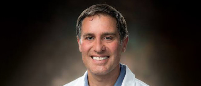 Professional headshot of a smiling man in a white coat and blue shirt