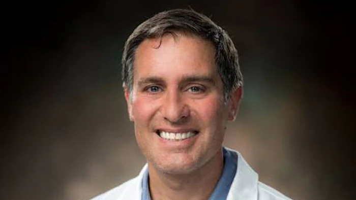 Professional headshot of a smiling man in a white coat and blue shirt