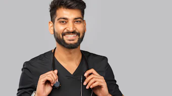 Professional headshot of a smiling man in black v-neck shirt holding a stethoscope