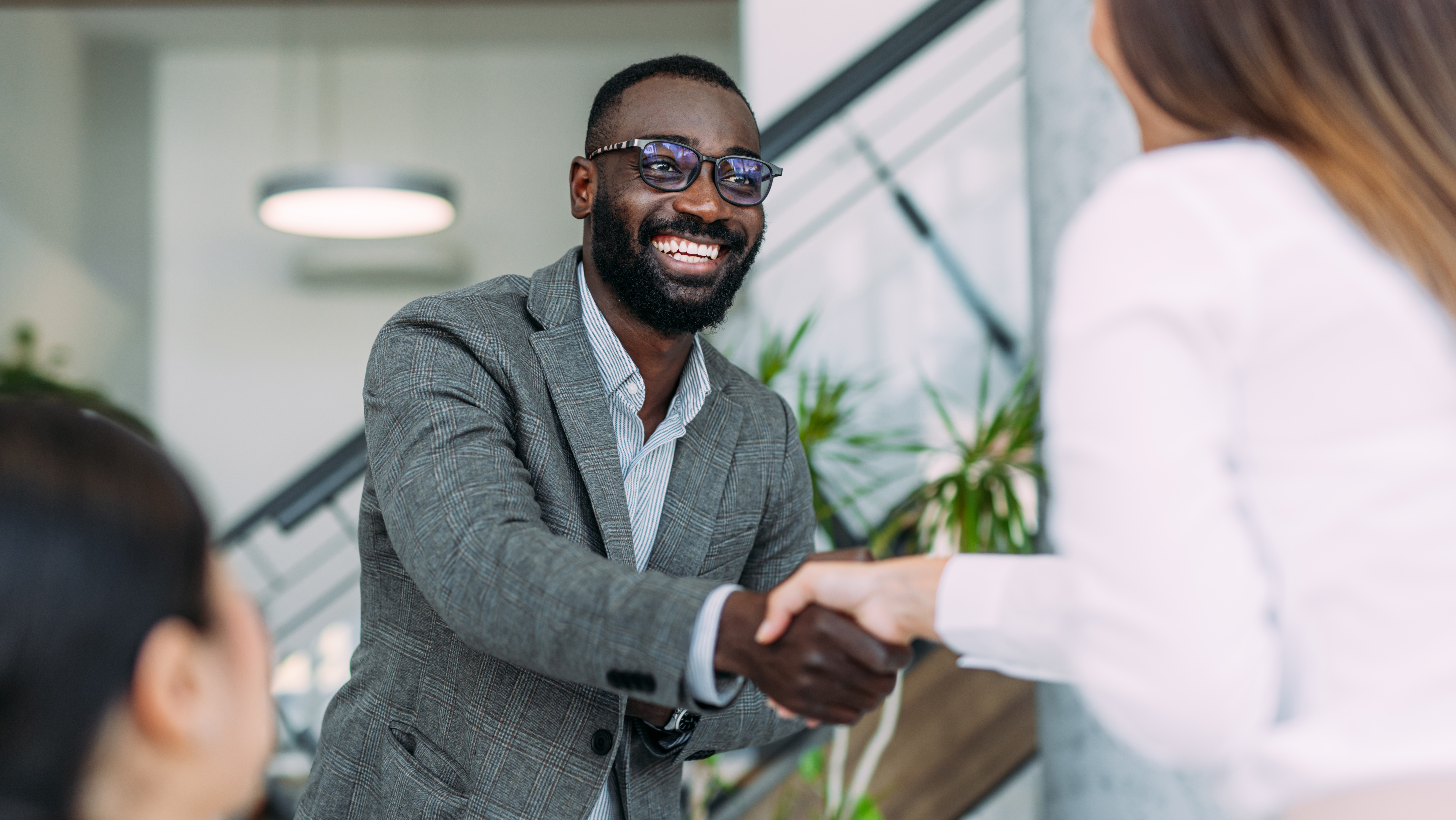 Professional man in gray blazer and glasses shaking hands with woman in modern office with plants and natural lighting