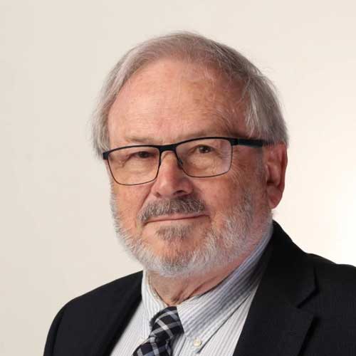 Professional headshot of an older man wearing glasses, striped shirt, and dark blazer with plaid tie