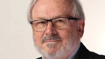 Professional headshot of an older man wearing glasses, striped shirt, and dark blazer with plaid tie